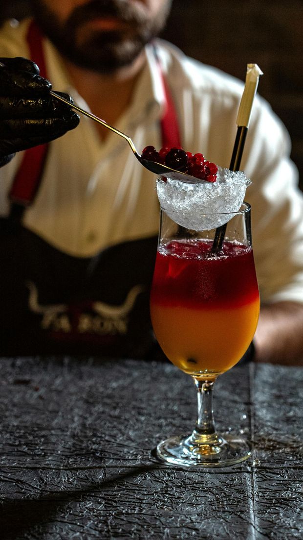 A bartender in a black apron uses a spoon to add red berries to an iced, layered orange and red cocktail in a glass.