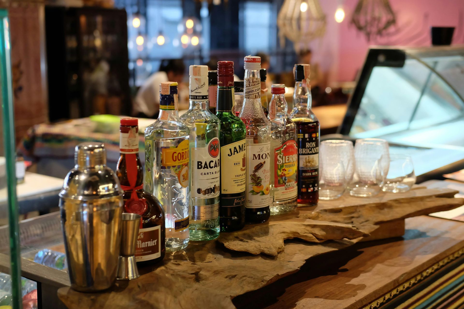 A wooden counter displays a metal cocktail shaker, various spirit bottles, and two empty glasses in a dimly lit bar.