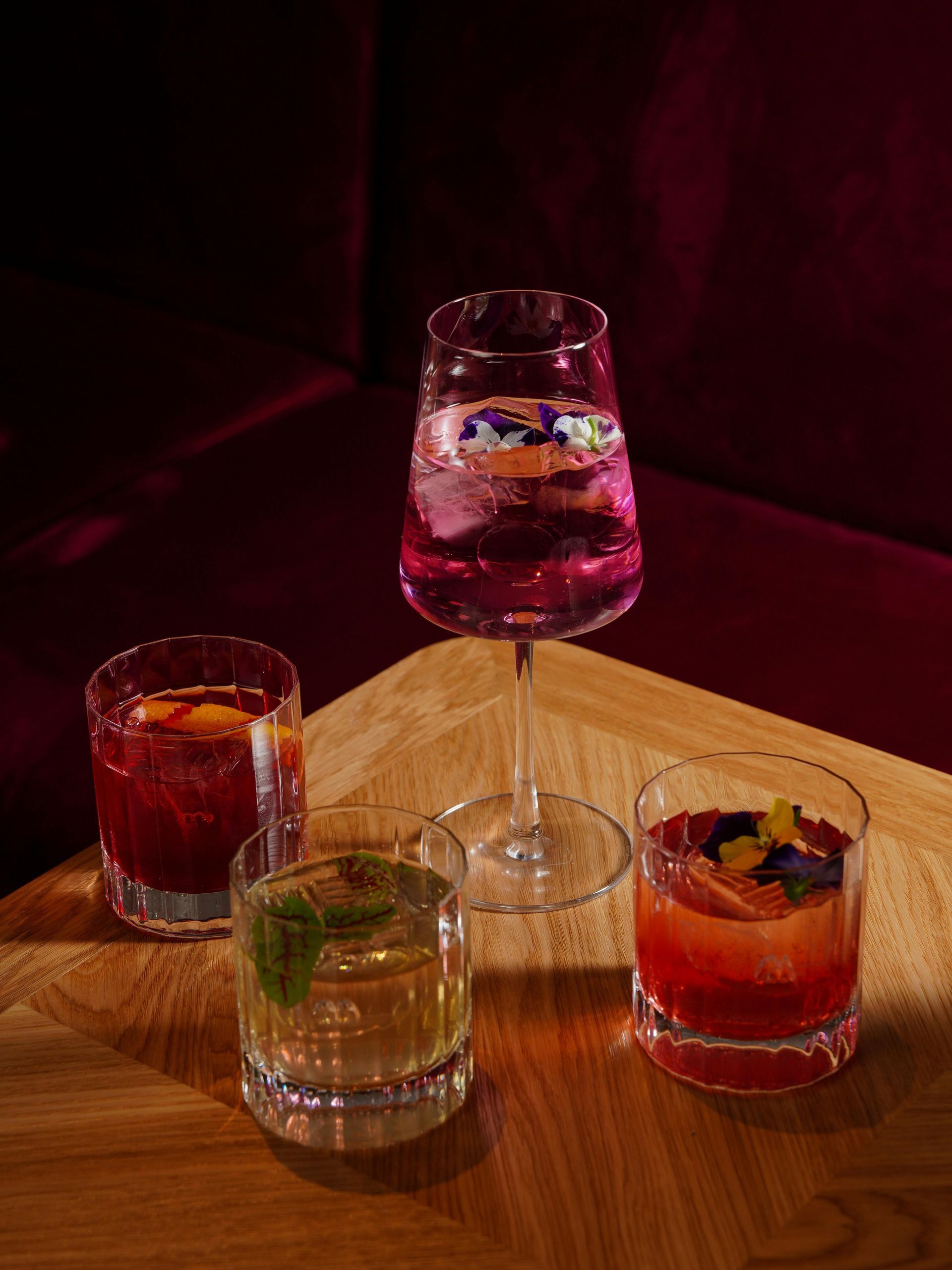 Four colorful cocktails in various glasses sit on a wooden table against a dark, moody background.