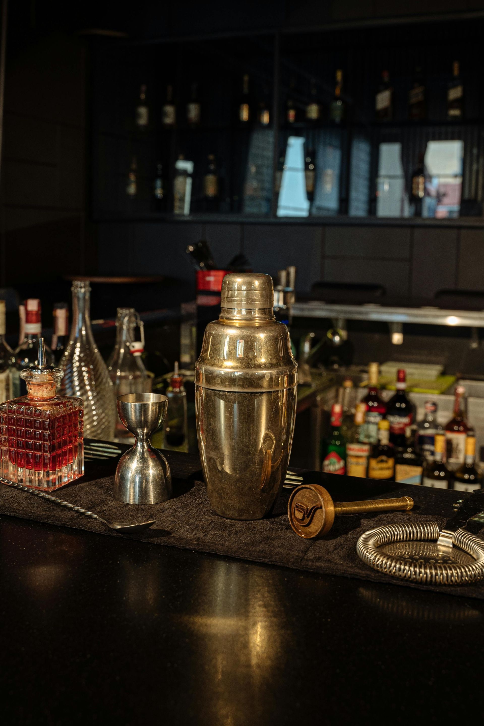 A stainless steel cocktail shaker, jigger, and bar tools on a dark bar mat, with shelves of liquor bottles in the background.