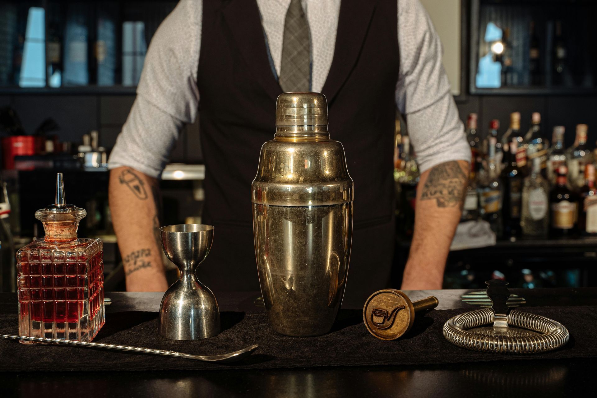 A bartender stands behind a bar with a cocktail shaker, a jigger, a mixing spoon, and a glass bottle filled with red liquid.