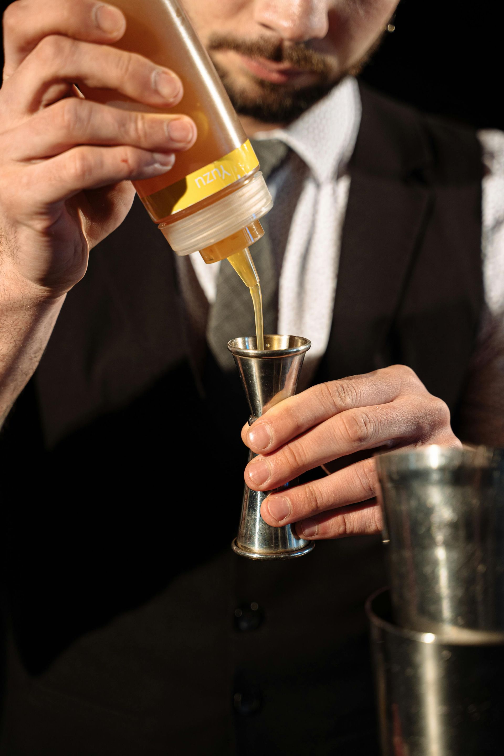 A bartender in a vest and tie pours golden liquid from a plastic squeeze bottle into a metal measuring jigger.