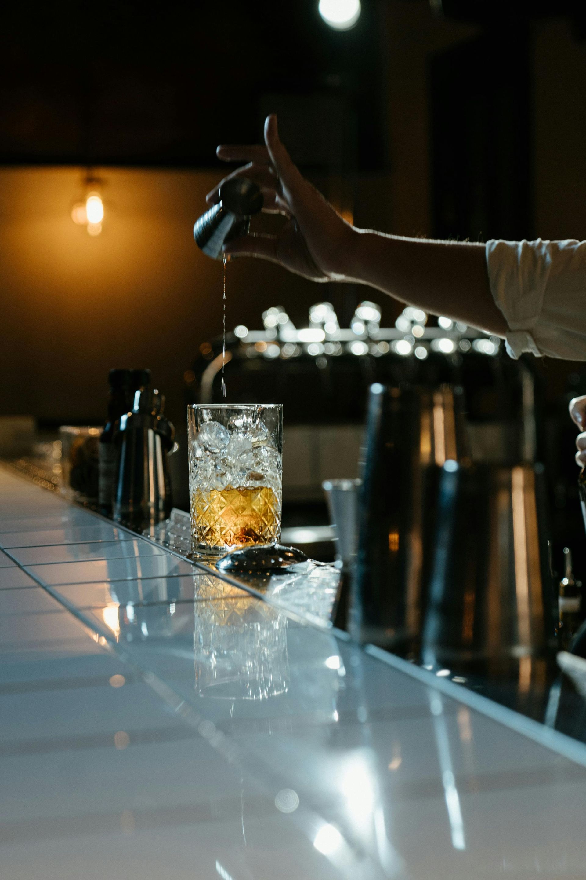 A person pours a measured drink into a glass filled with ice on a reflective, white tiled bar counter.