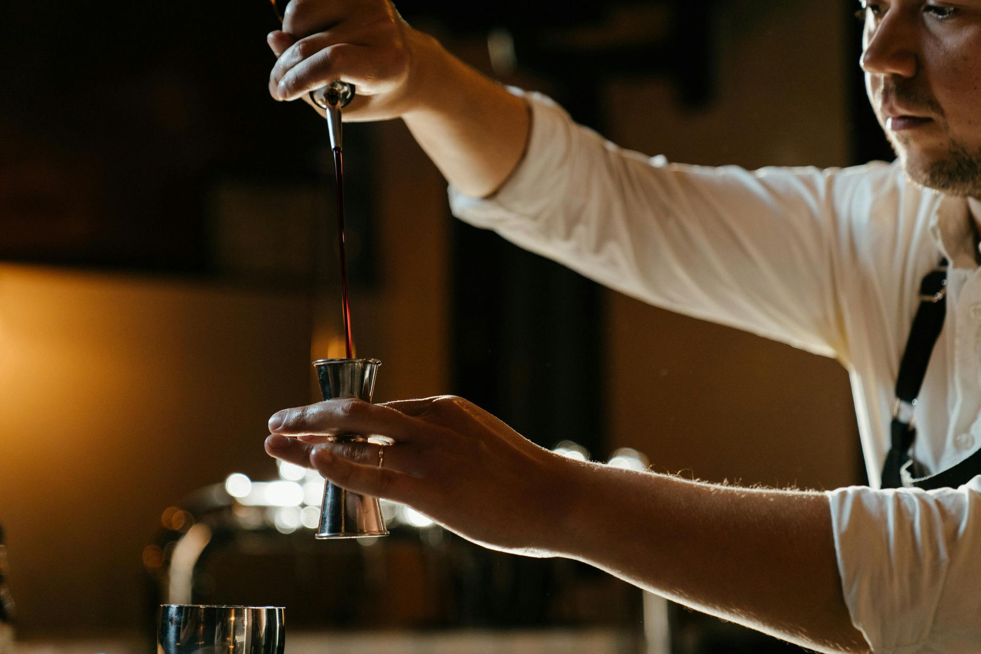 A bartender pours a dark liquid from a bottle into a metal jigger at a bar.