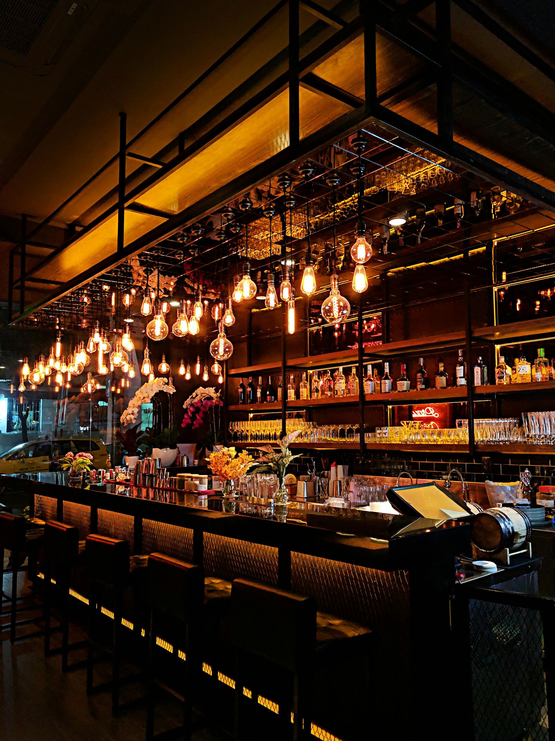 A dimly lit bar with rows of hanging Edison bulbs over a long counter and shelves stocked with various liquor bottles.