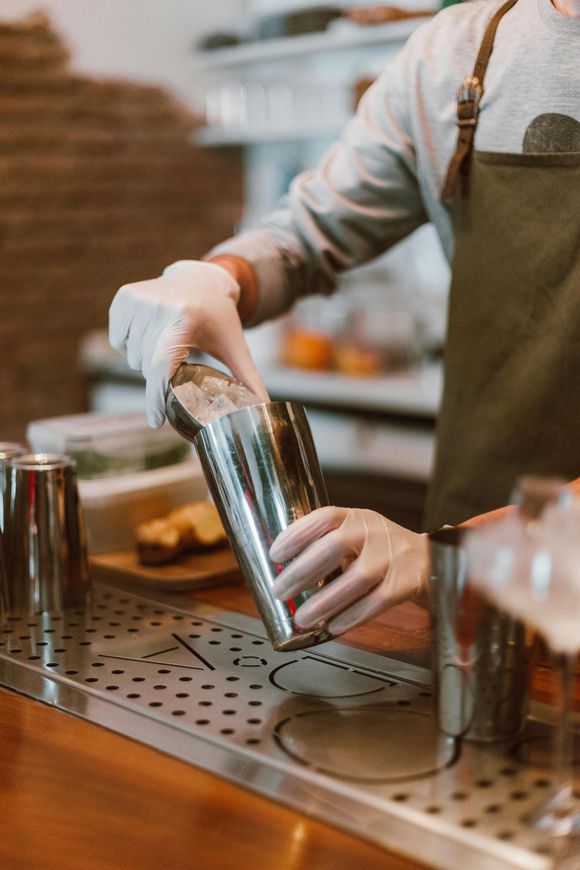 A person wearing an apron and gloves drops ice into a stainless steel cocktail shaker on a metal bar mat.
