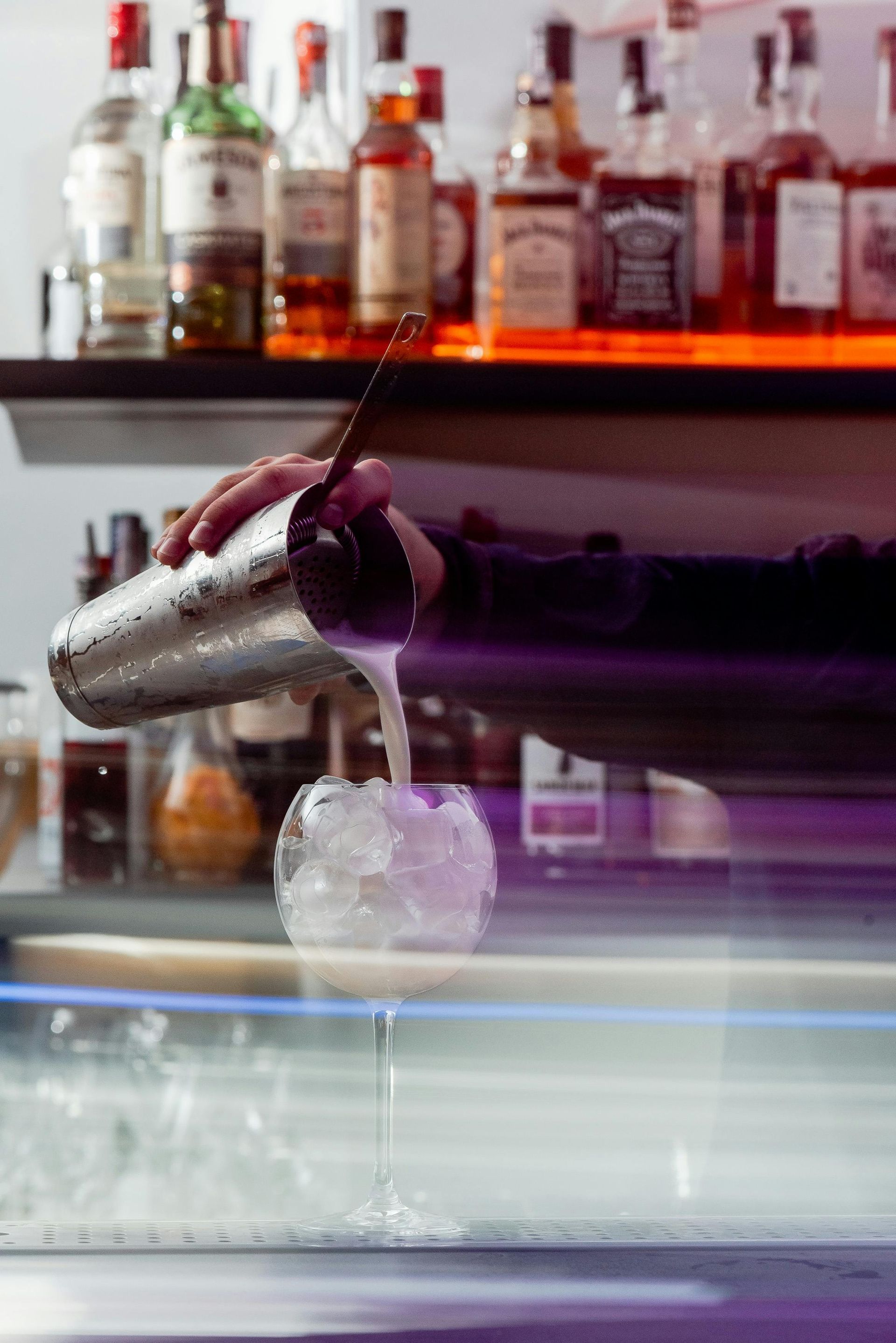 A bartender pours a cocktail from a metal shaker into a glass filled with ice at a dimly lit bar.