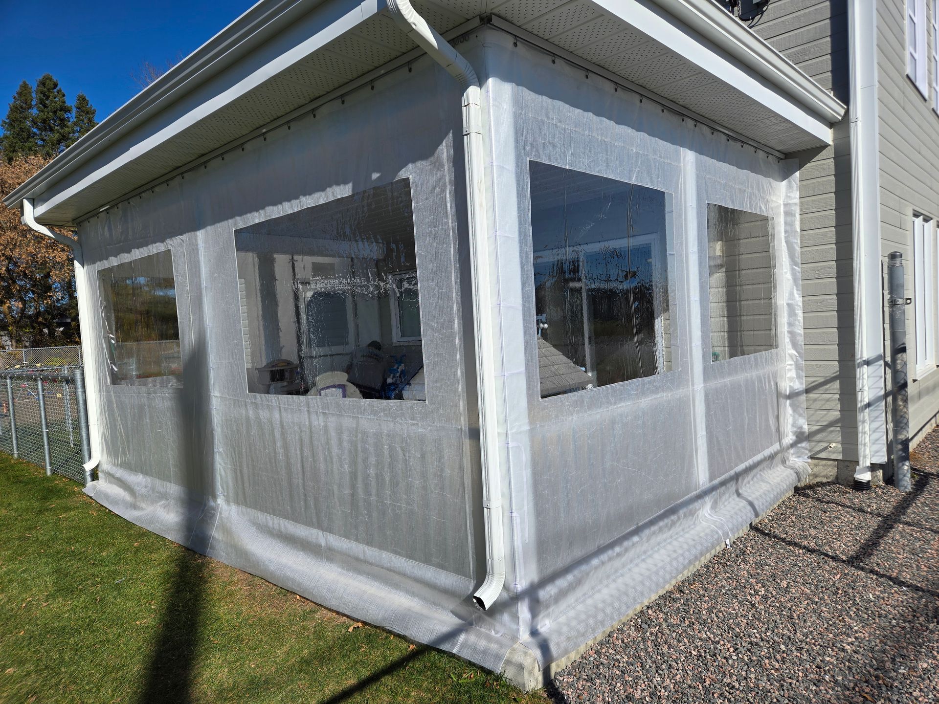 Véranda fermée avec fenêtres en vinyle transparent, attenante à une maison avec pelouse verte et gravier.