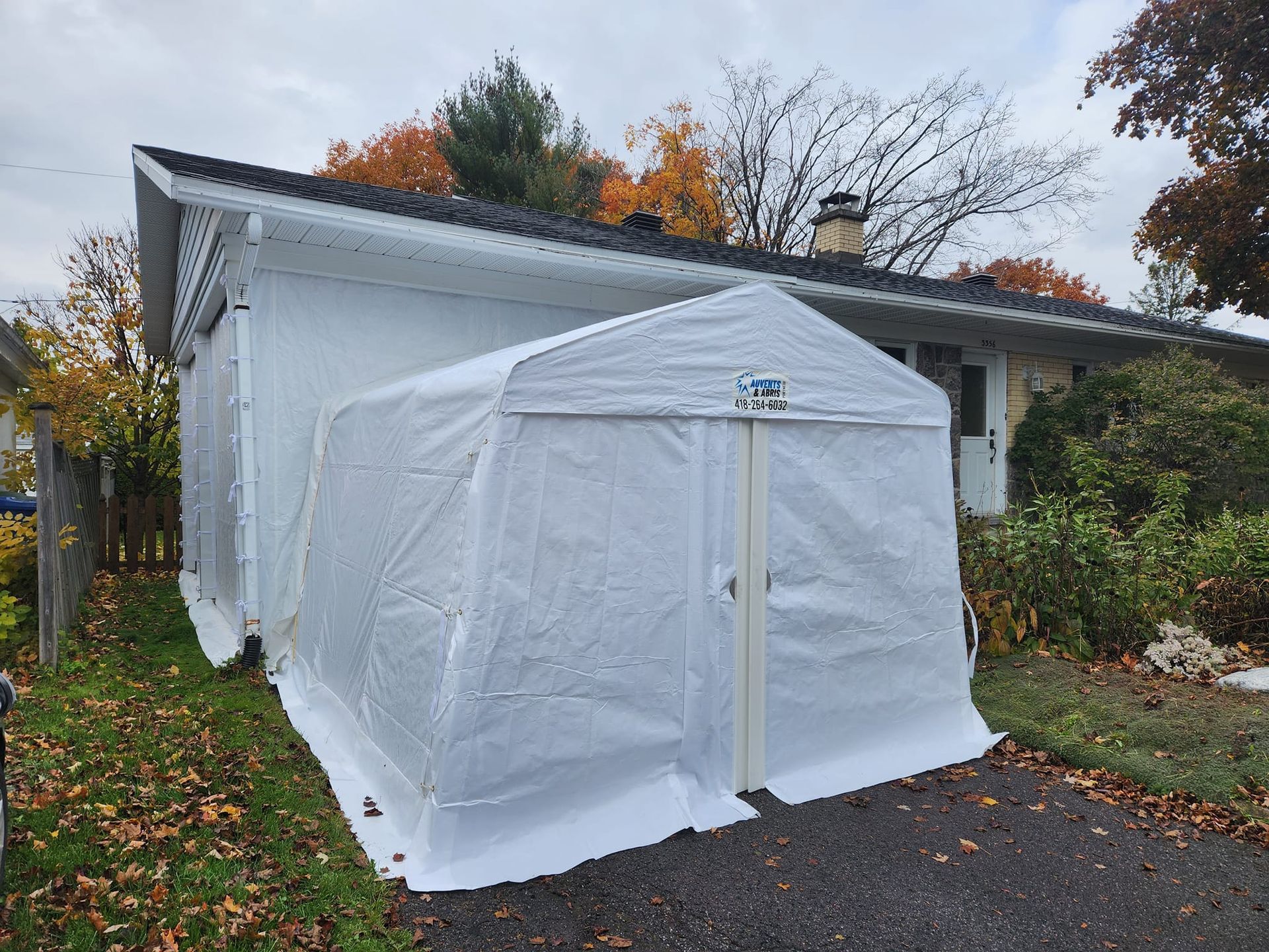 Abri en bâche blanche adossé à une maison blanche avec allée et feuillage d'automne.