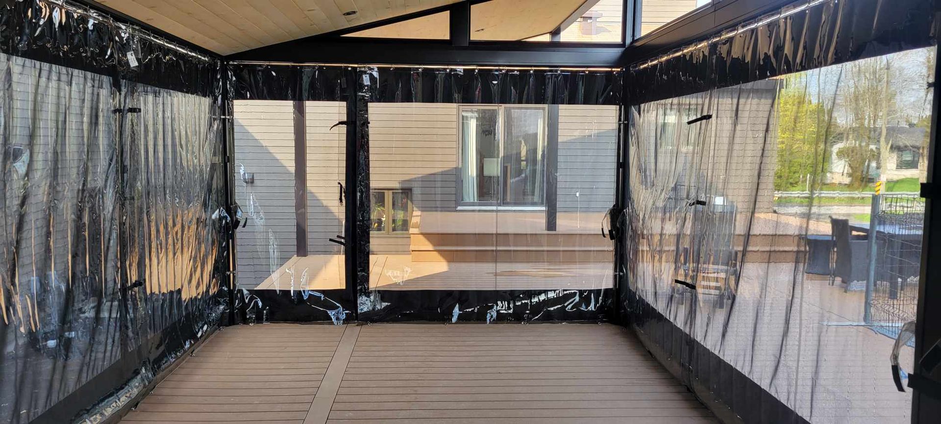 Espace extérieur clos avec rideaux en vinyle transparent, parquet et finitions noires. Vue sur une terrasse et des arbres.