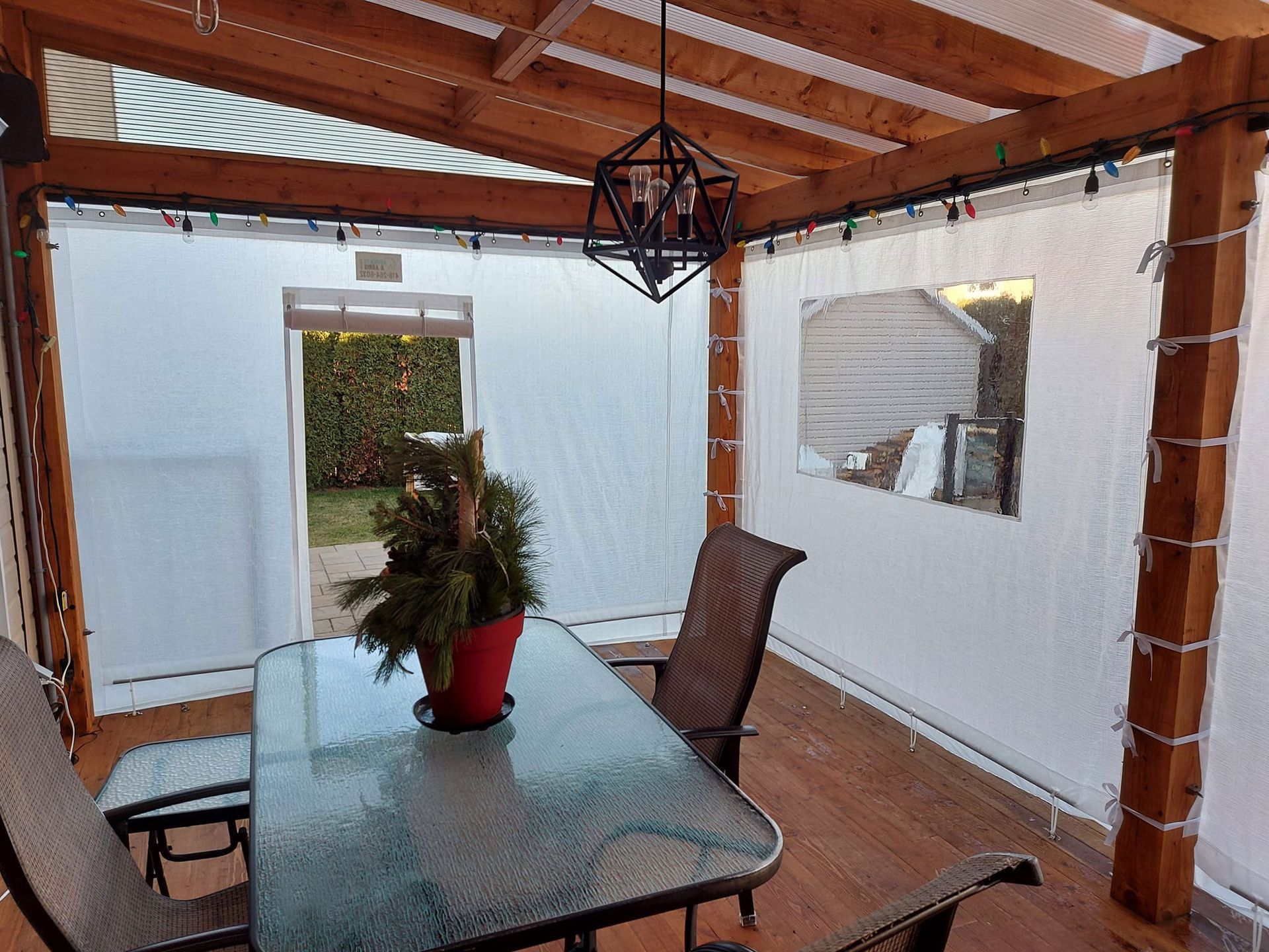 Patio clos avec table en verre, chaises et suspension lumineuse. Rideaux blancs, plantes vertes et poutres apparentes.
