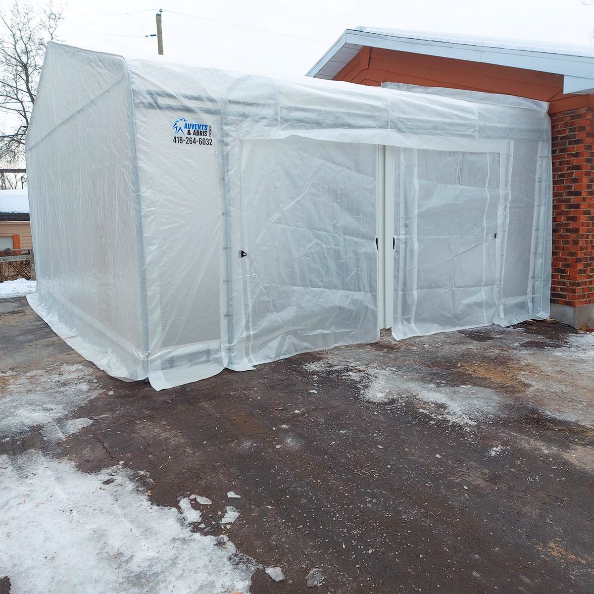 Un abri en plastique transparent à deux portes se trouve à côté d'un bâtiment en briques. Le sol est recouvert de neige.