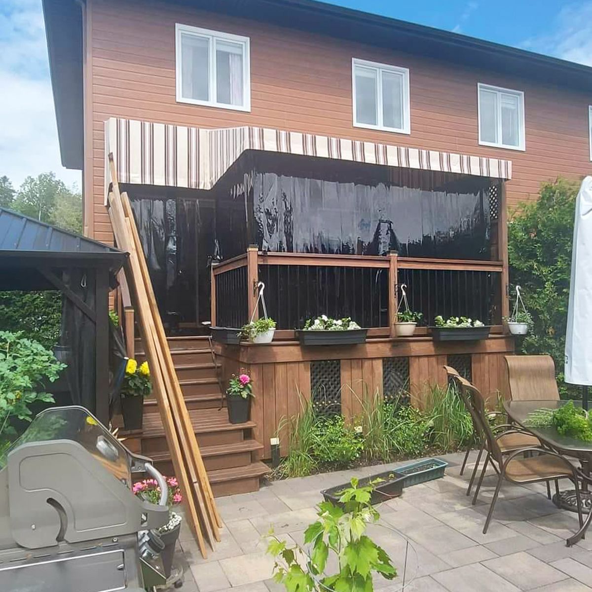 Terrasse arrière avec auvent, rideaux d'intimité, jardinières et barbecue. Revêtement extérieur de la maison en bois brun.