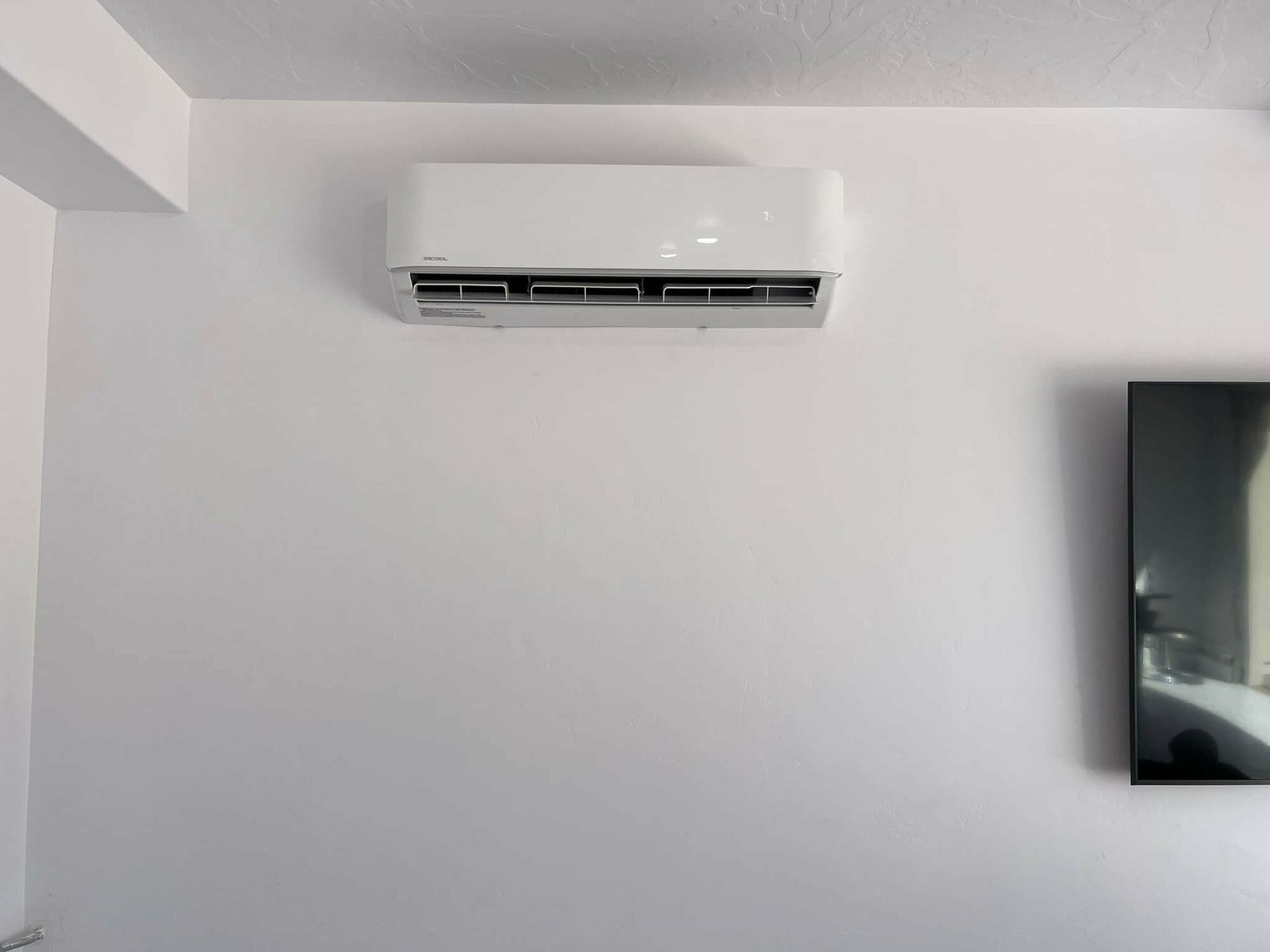 A white air conditioner is mounted on a white wall next to a flat screen tv.