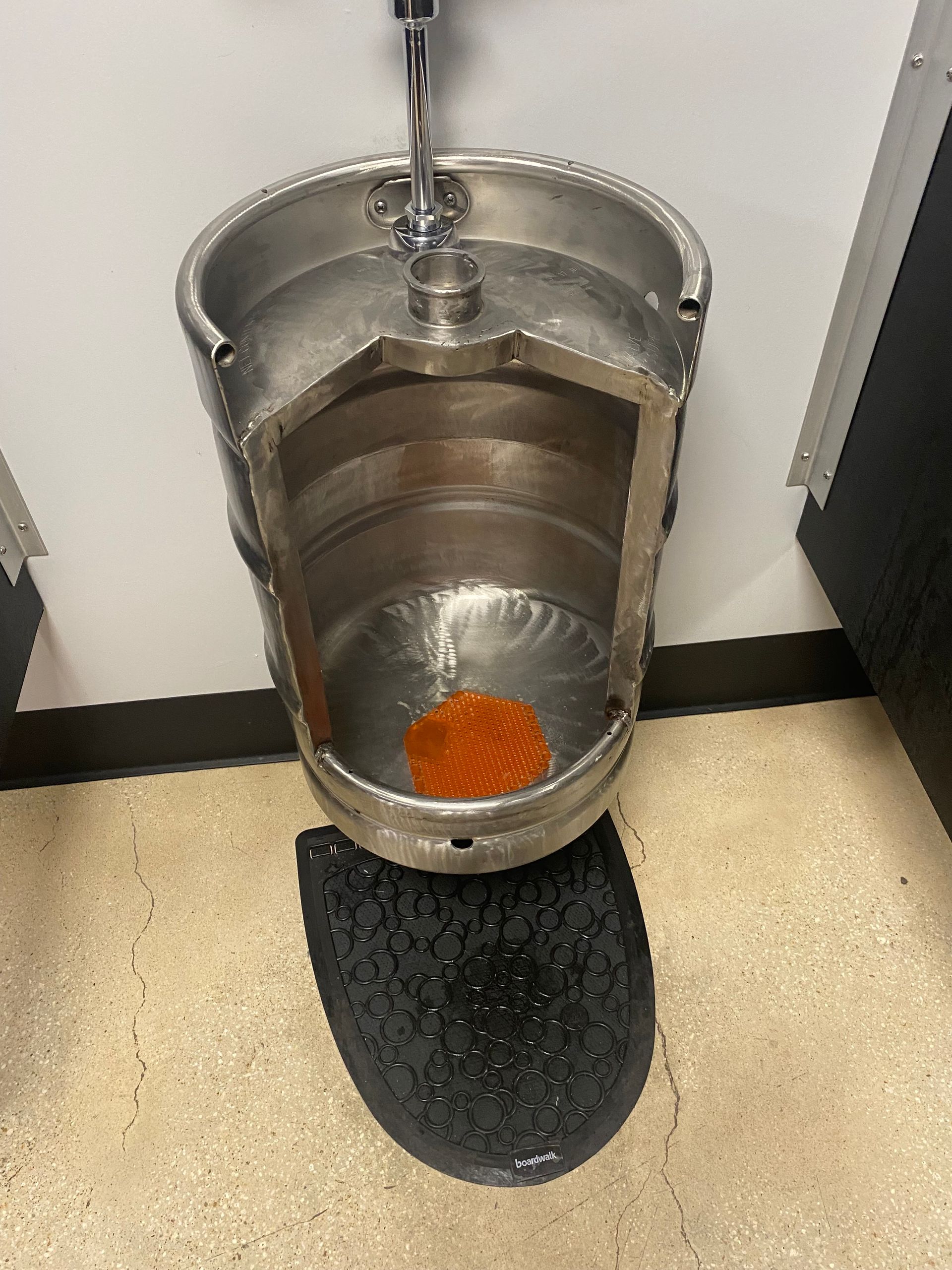 A silver beer keg converted into a wall-mounted urinal with a black floor mat in a restroom.