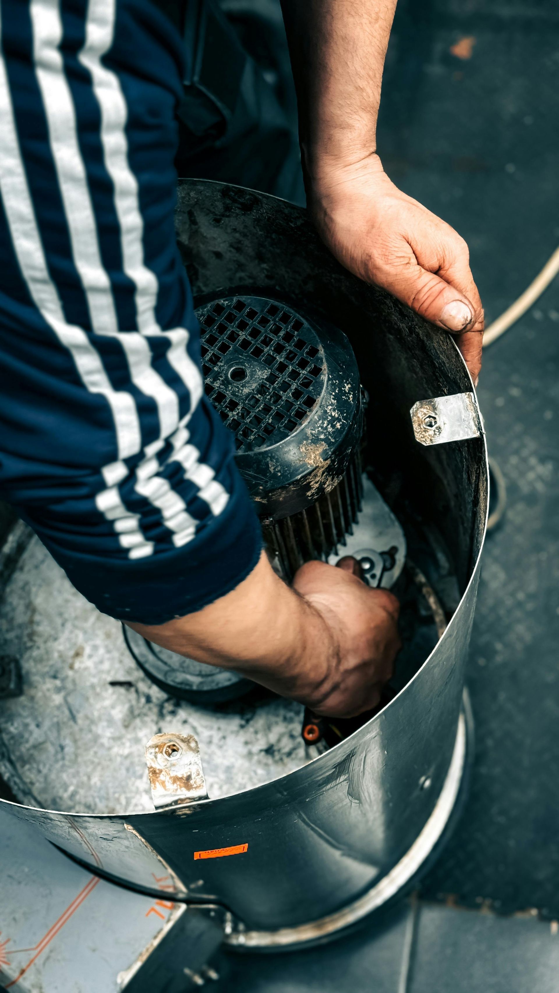 A person in a blue Adidas jacket reaches into a cylindrical metal canister to adjust or clean a motor component inside.