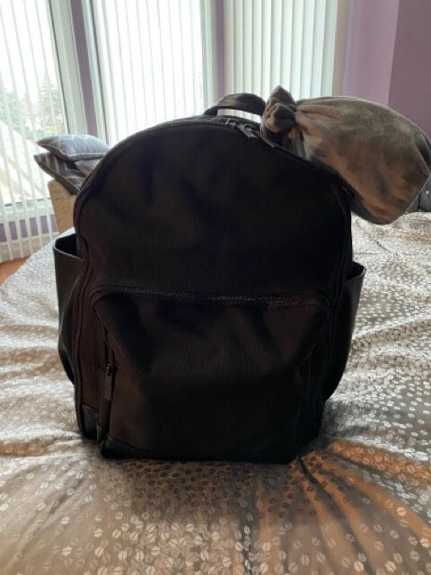 beis the backpack black backpack fully packed on gray bedspread gray travel pillow attached windows in background
