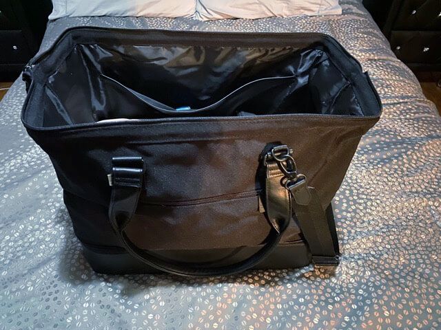 beis the convertible weekender black weekender bag open top empty bag on gray bedspread black leather handle black carrying strap zippered compartment