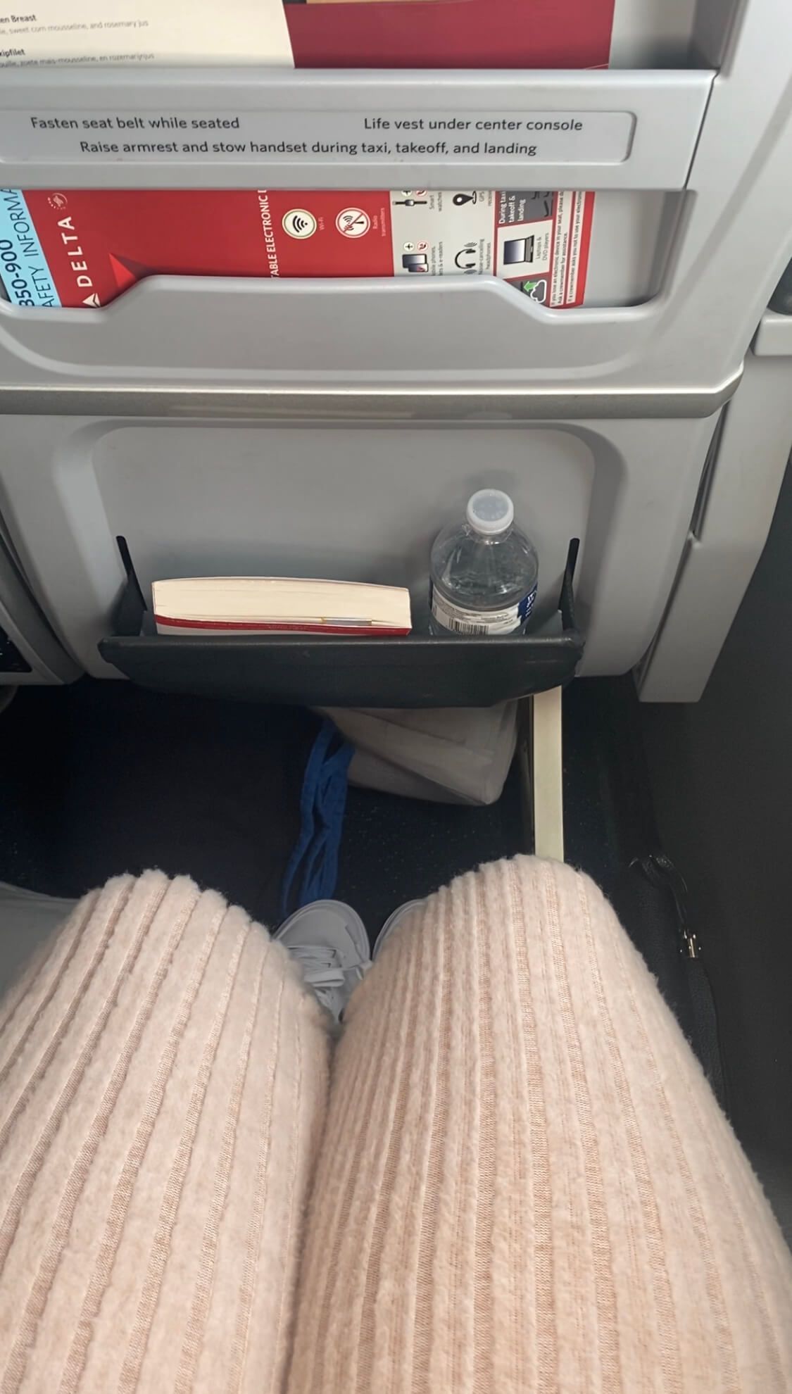 delta premium select seat legroom tan pants white shoes water bottle and book in seat pocket showing several inches of legroom