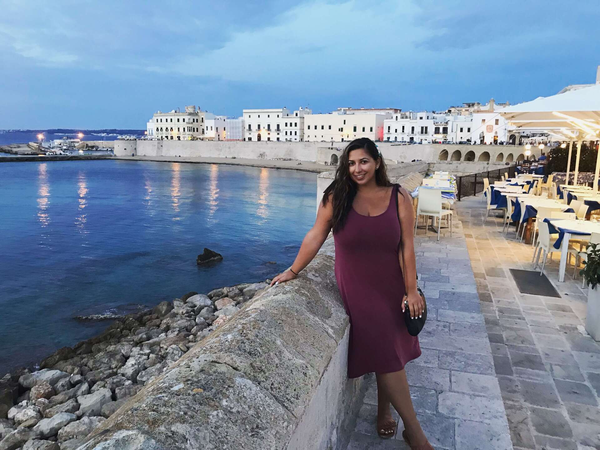 woman dark hair purple dress with Gallipoli city in background white buildings fortress sea at sunset