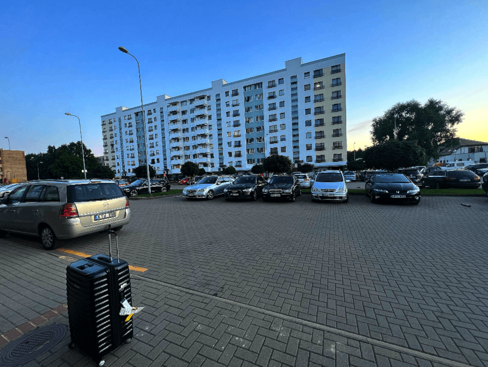 black samsonite checked luggage white luggage tags white apartment building and parking lot with multiple cars in Lithuania