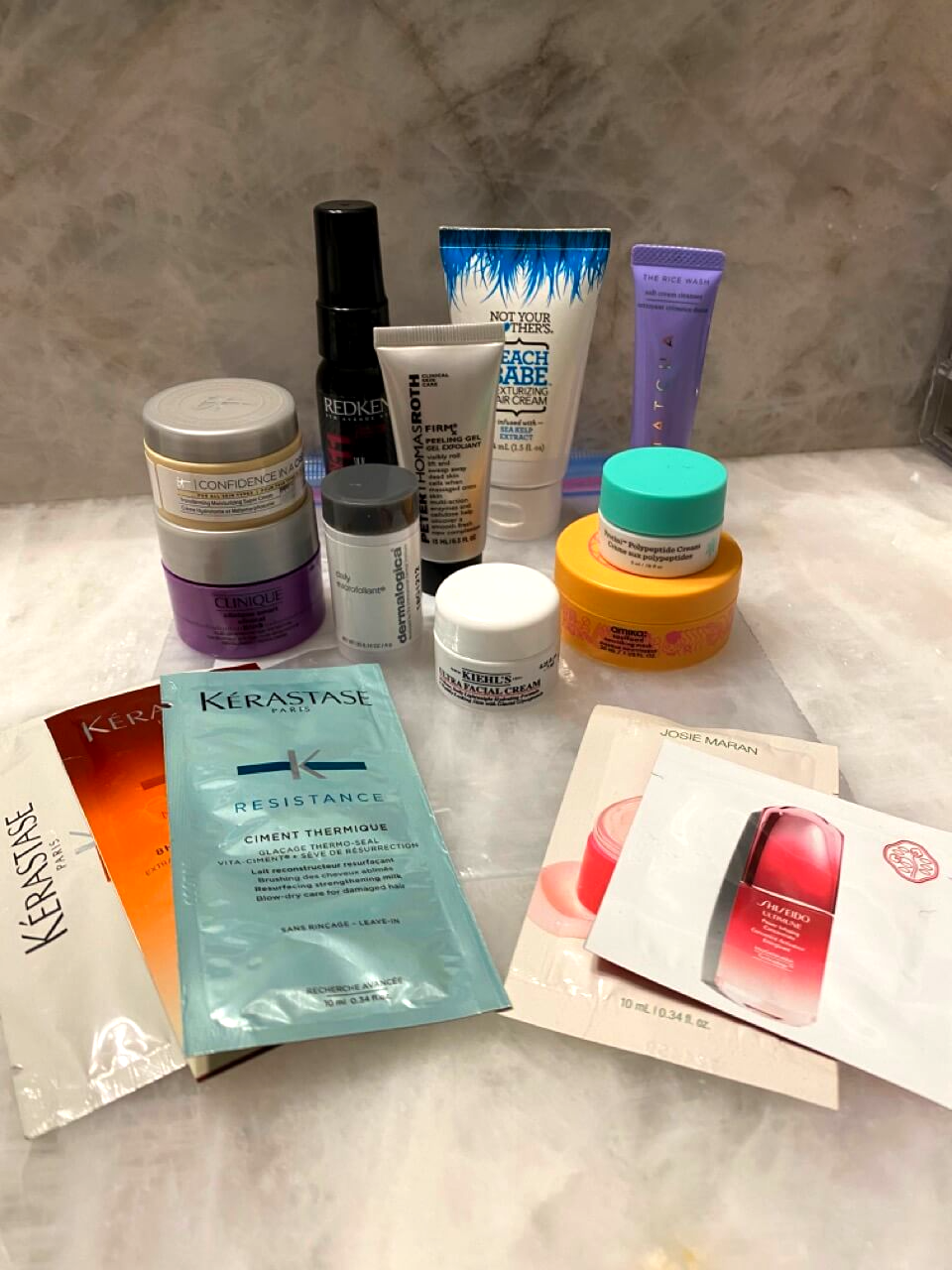 various toiletries on white marble counter top five packets of hair and facial serums and ten mini containers of facial cream, hair styling tools, and face washes in assorted colors