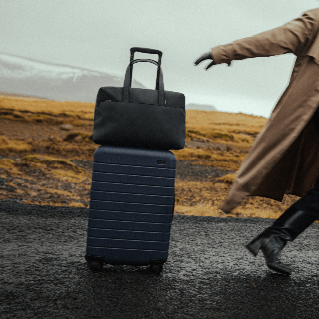 away carry on suitcase blue and black bag on suitcase on asphalt person with tan trench coat