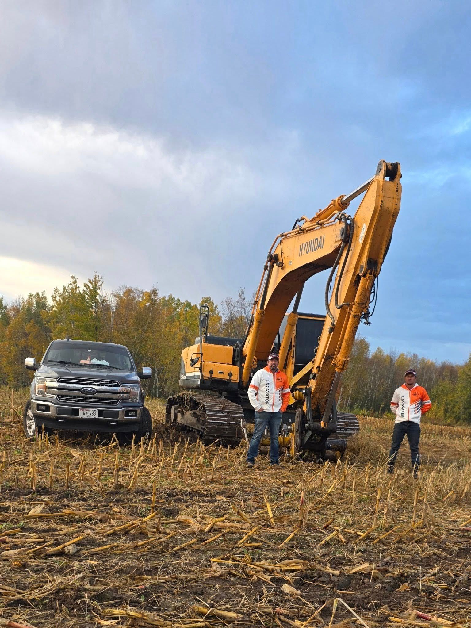 Two people stand in a harvested field next to a large yellow excavator and a dark pickup truck under a blue sky.