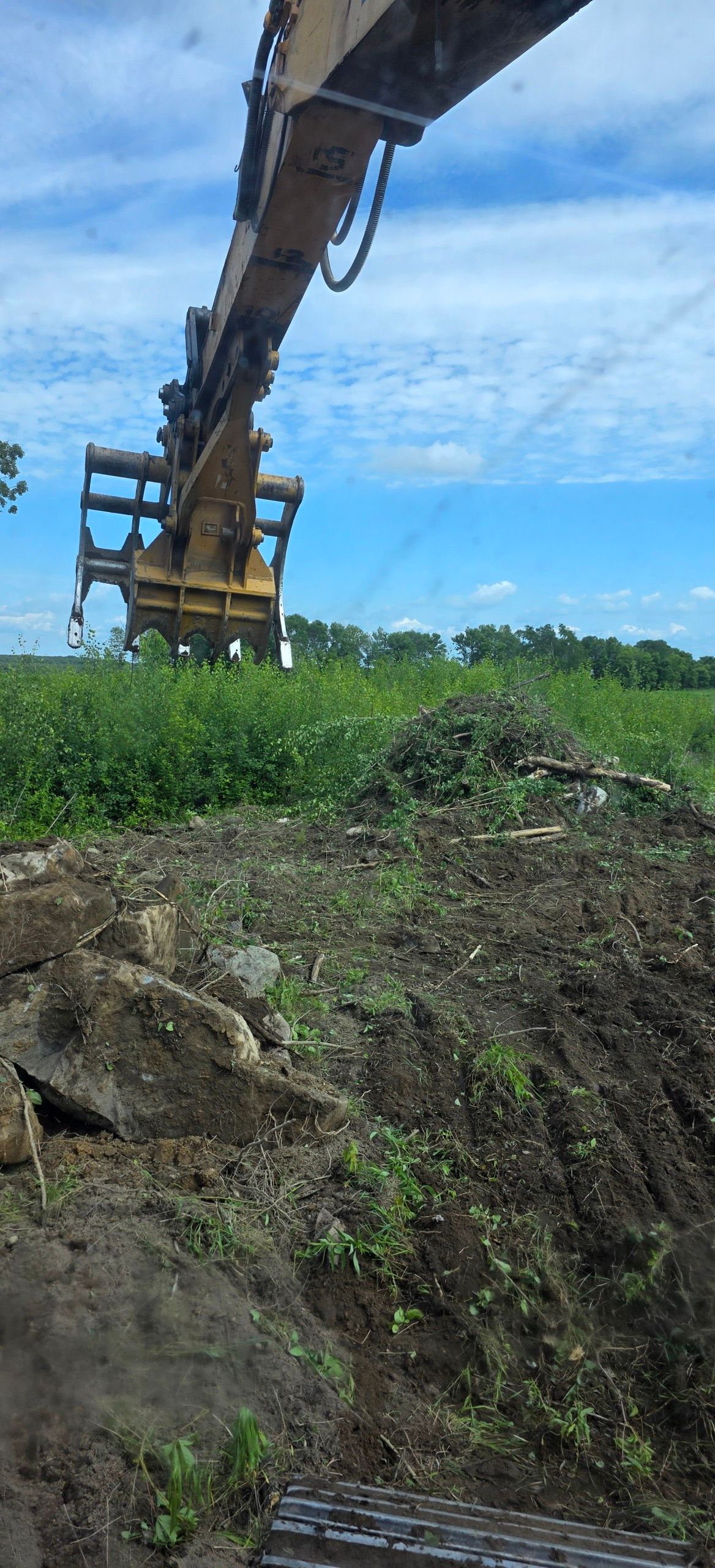 A yellow excavator arm with a grapple attachment extends over a mound of dirt and vegetation under a bright blue sky.