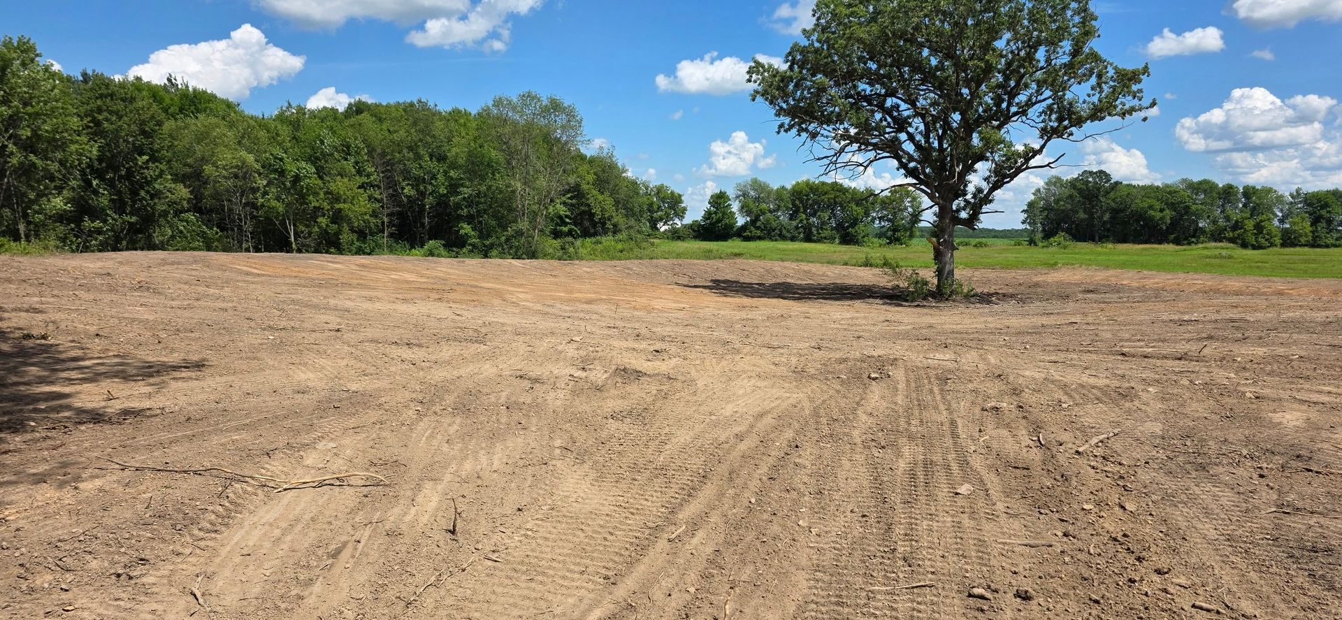 A large, cleared field of dry, brown soil under a sunny blue sky with clouds, featuring a single tree in the distance.