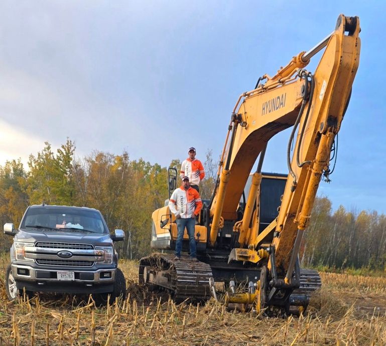 Two people standing on a yellow excavator parked next to a gray pickup truck in a harvested field.