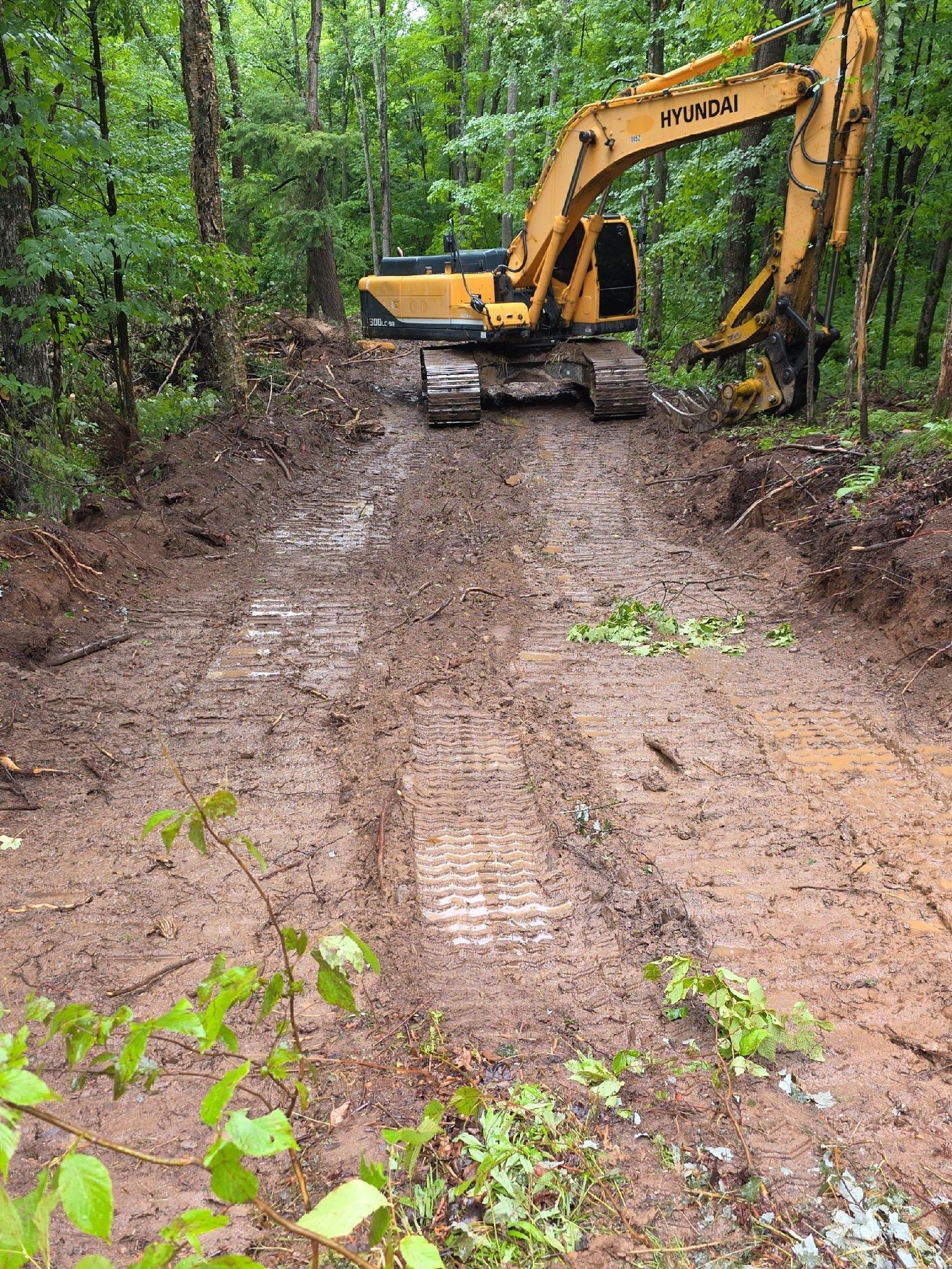 A yellow excavator parked on a muddy, rutted dirt road in a dense, green forest.