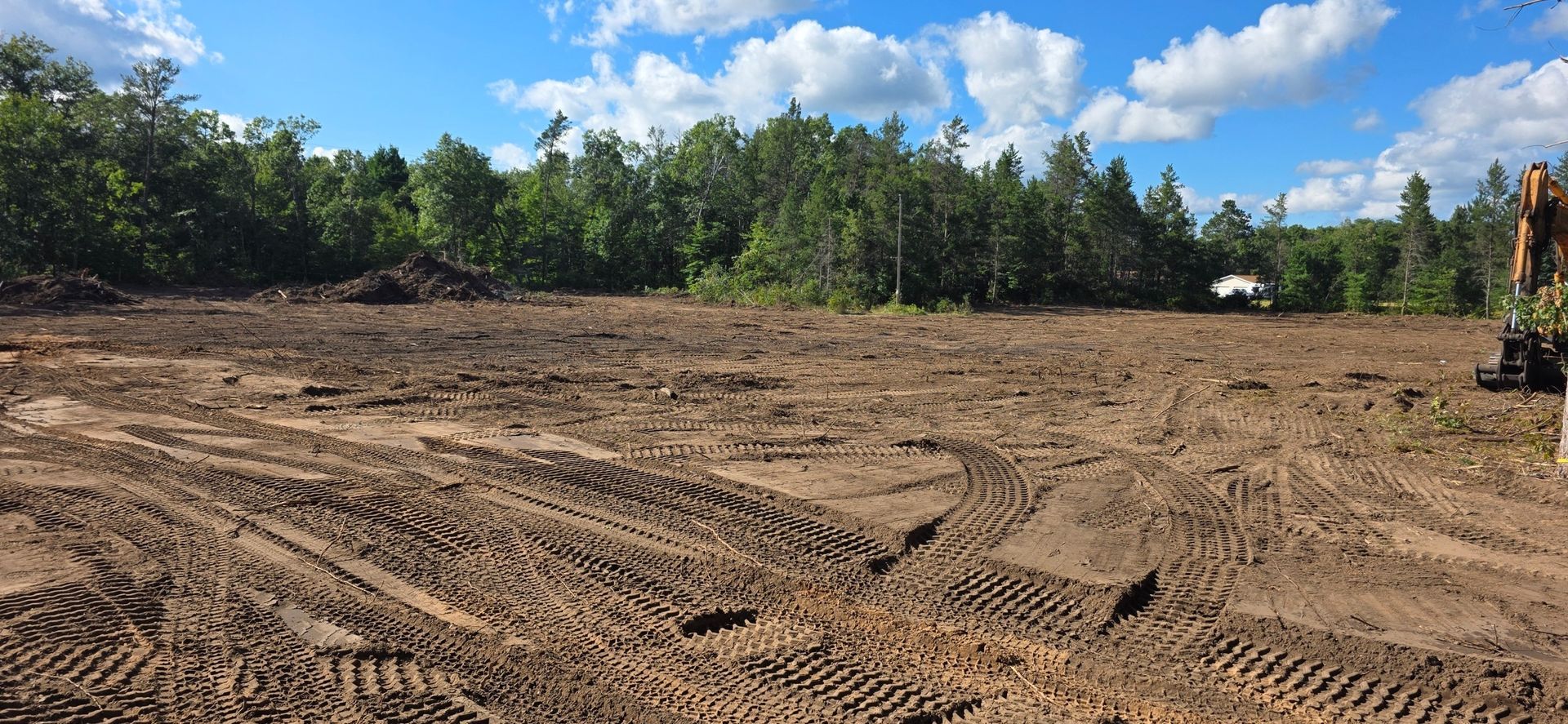 A cleared lot of brown dirt with tire tracks, bordered by a dense green forest under a blue sky with scattered clouds.