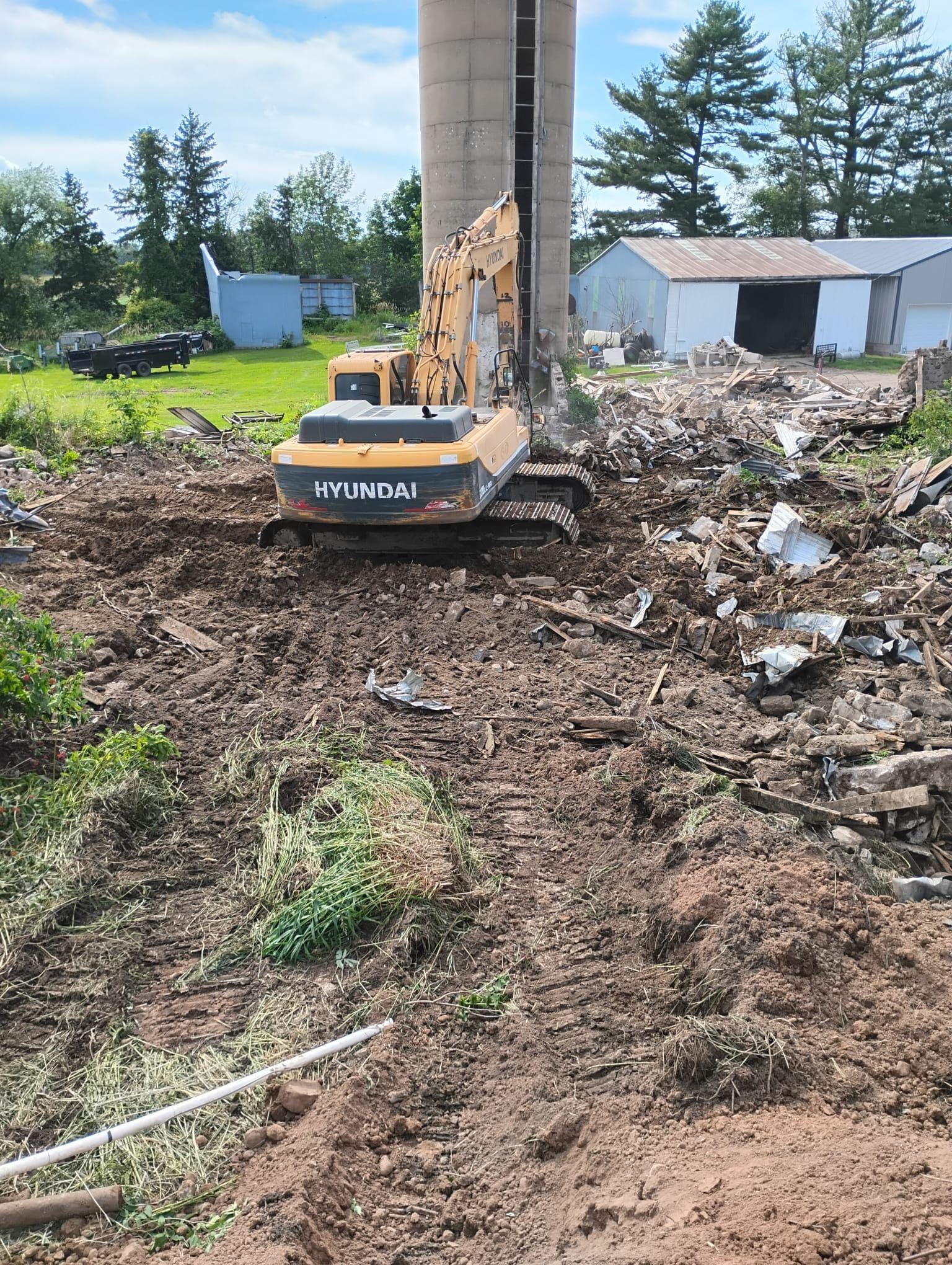 A yellow Hyundai excavator sits in a dirt clearing next to a concrete silo during a demolition project.