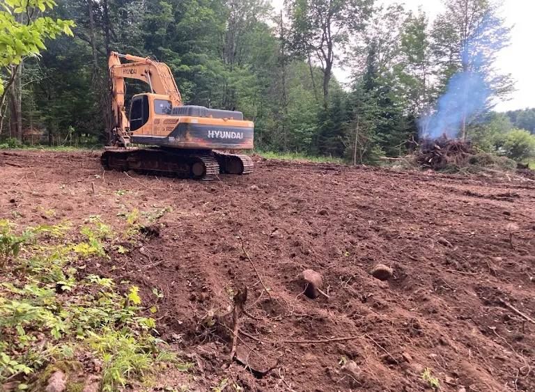 A yellow excavator parked on cleared brown earth with a smoldering brush pile in a forested area.