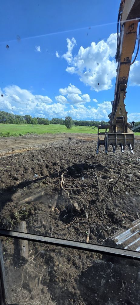 An excavator arm with a grapple attachment works on a muddy, cleared field under a bright blue, cloudy sky.