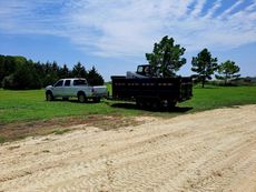 White pickup truck towing a black dump trailer on a grassy field under a blue sky.