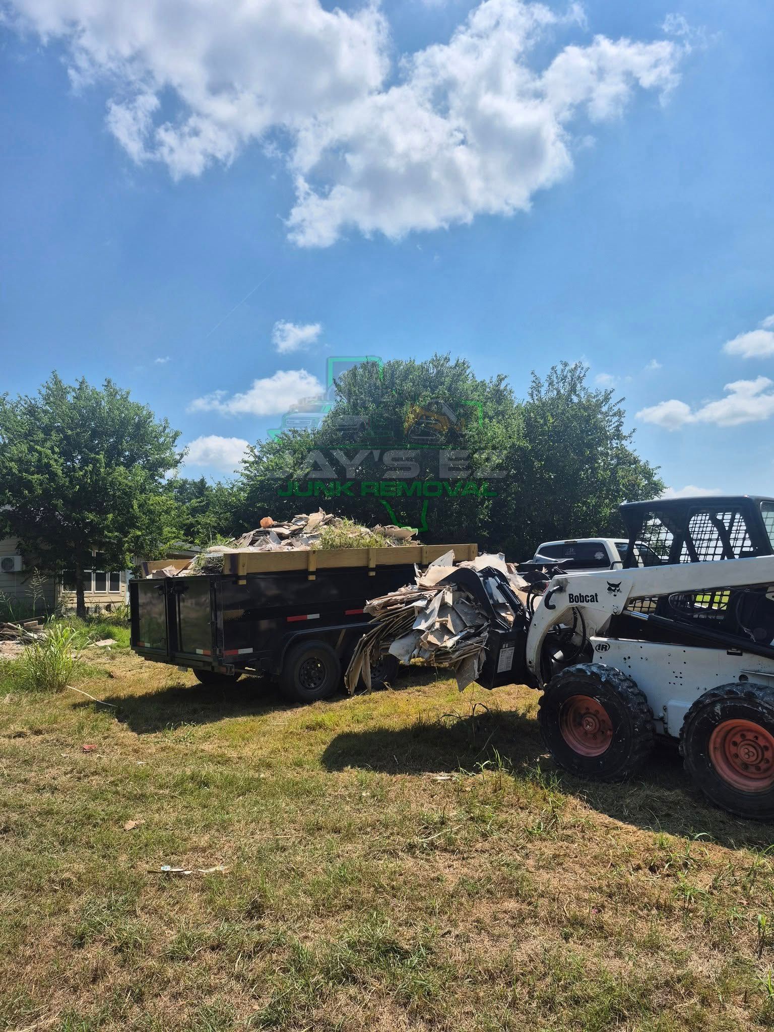 Bobcat loading debris into a trailer in a grassy area under a blue sky.