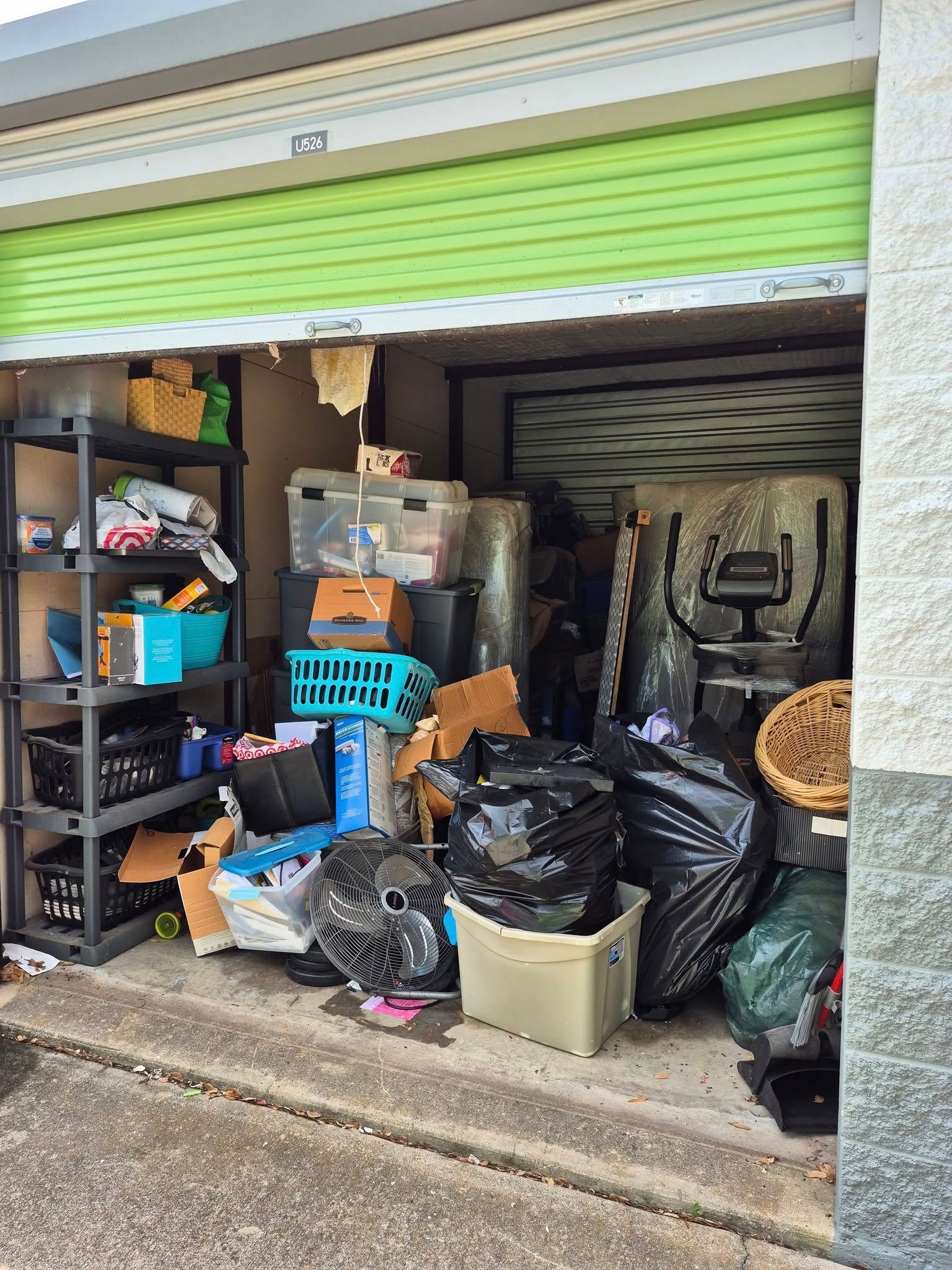 Storage unit overflowing with various items, including boxes, shelves, and trash bags. Green door is partially open.