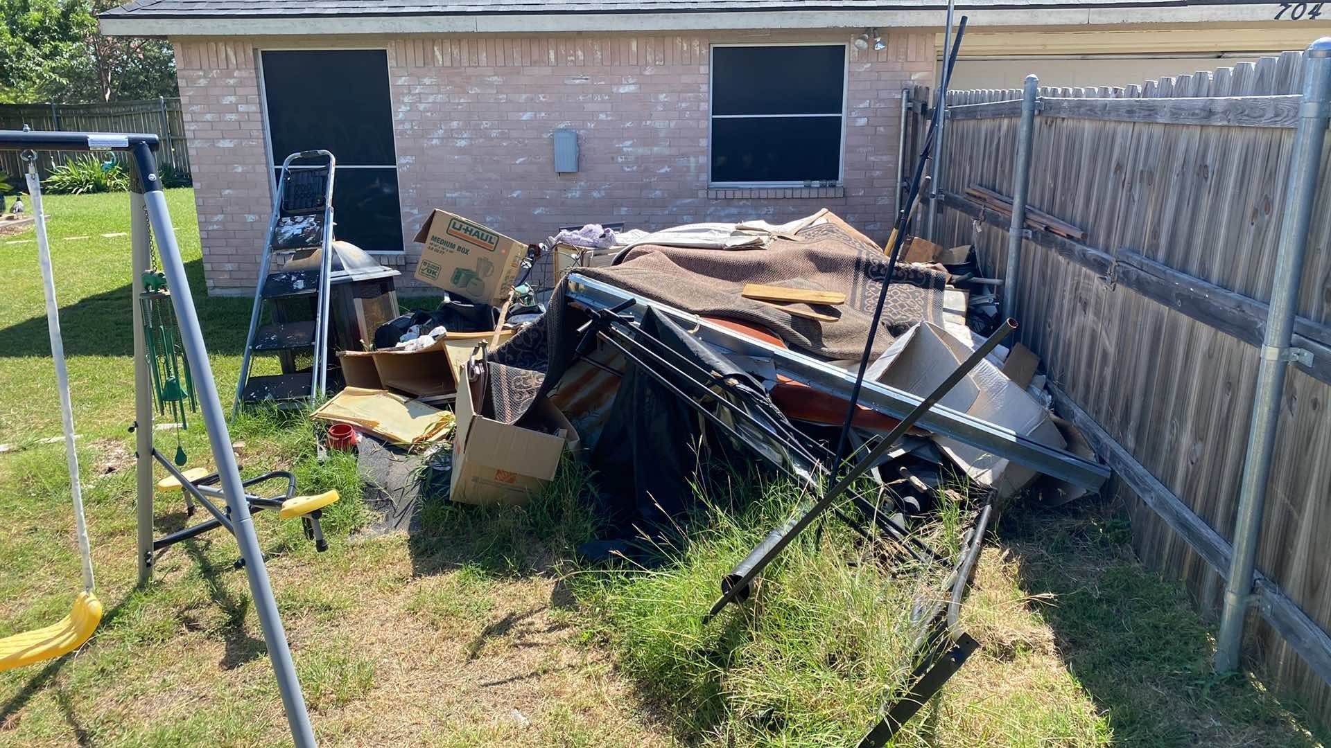 Pile of debris in a backyard, including cardboard boxes, metal, and wood near a fence and brick building.