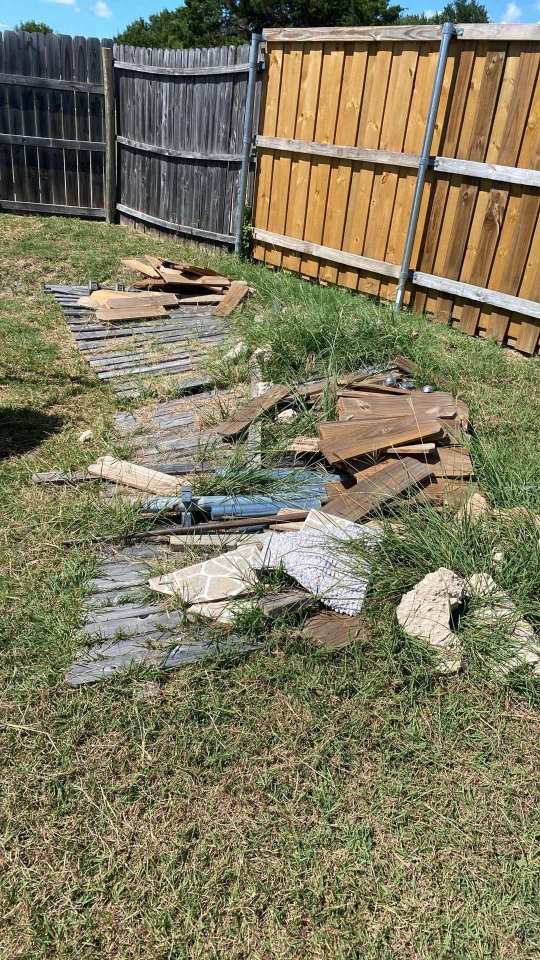 Debris of stone and wood in a grassy yard, next to weathered fences under a sunny sky.