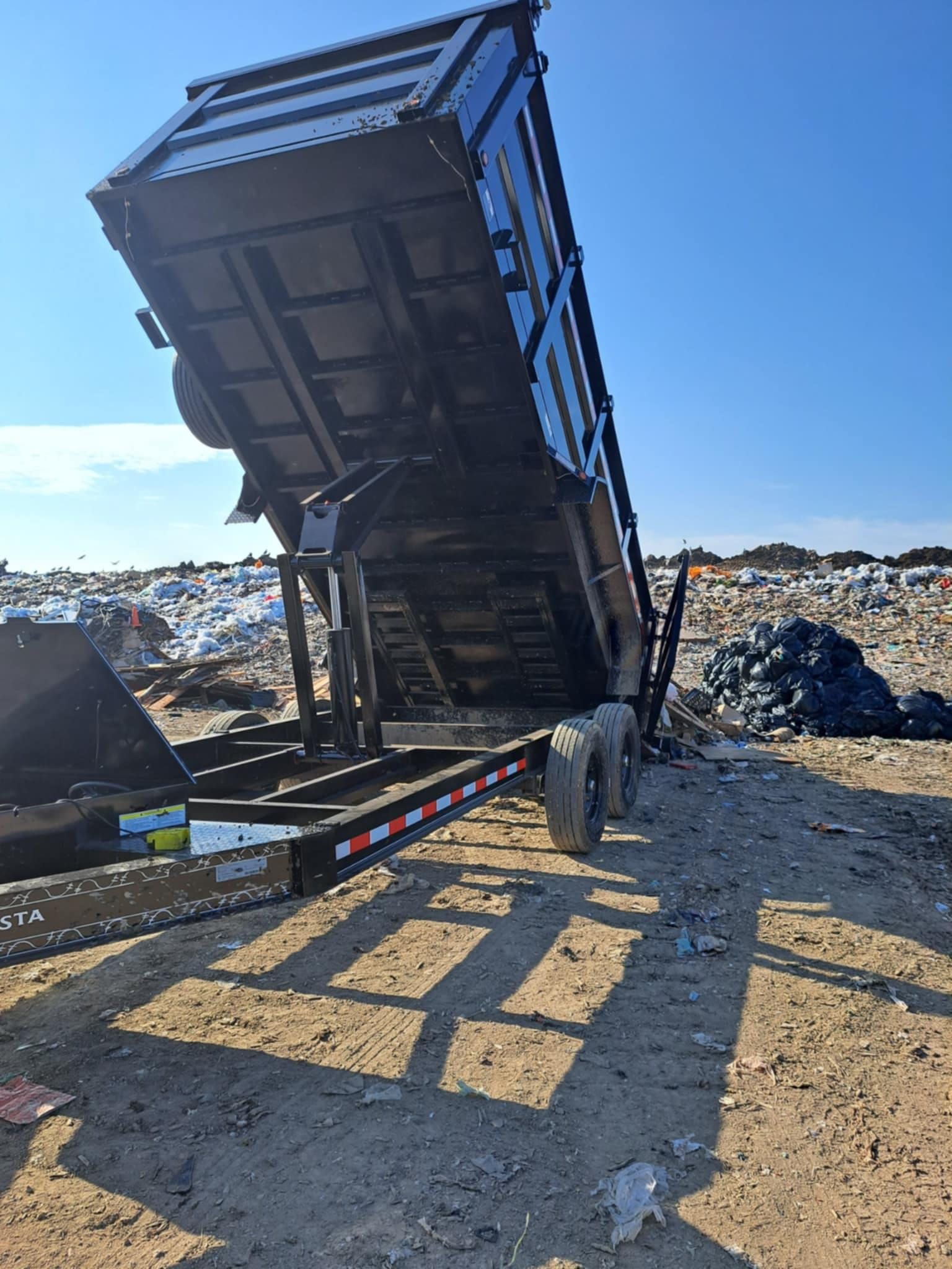 Black dump trailer raised, dumping trash at a landfill, sunny day.