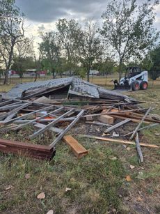Demolished shed with a Bobcat loader on a grassy field under a cloudy sky.