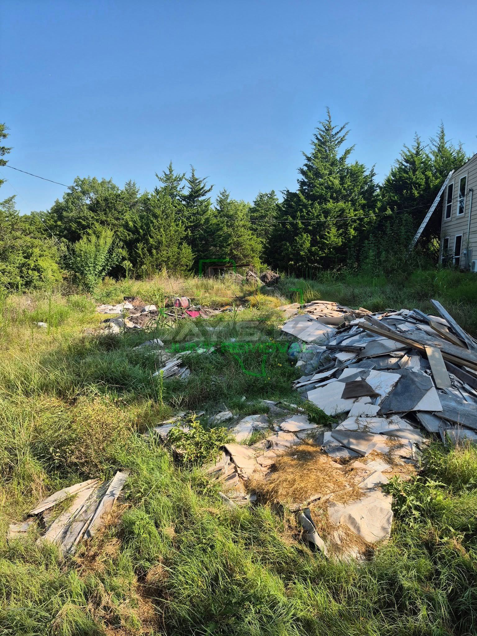 Debris pile on overgrown land under a blue sky, trees in the background, corner of building on right.