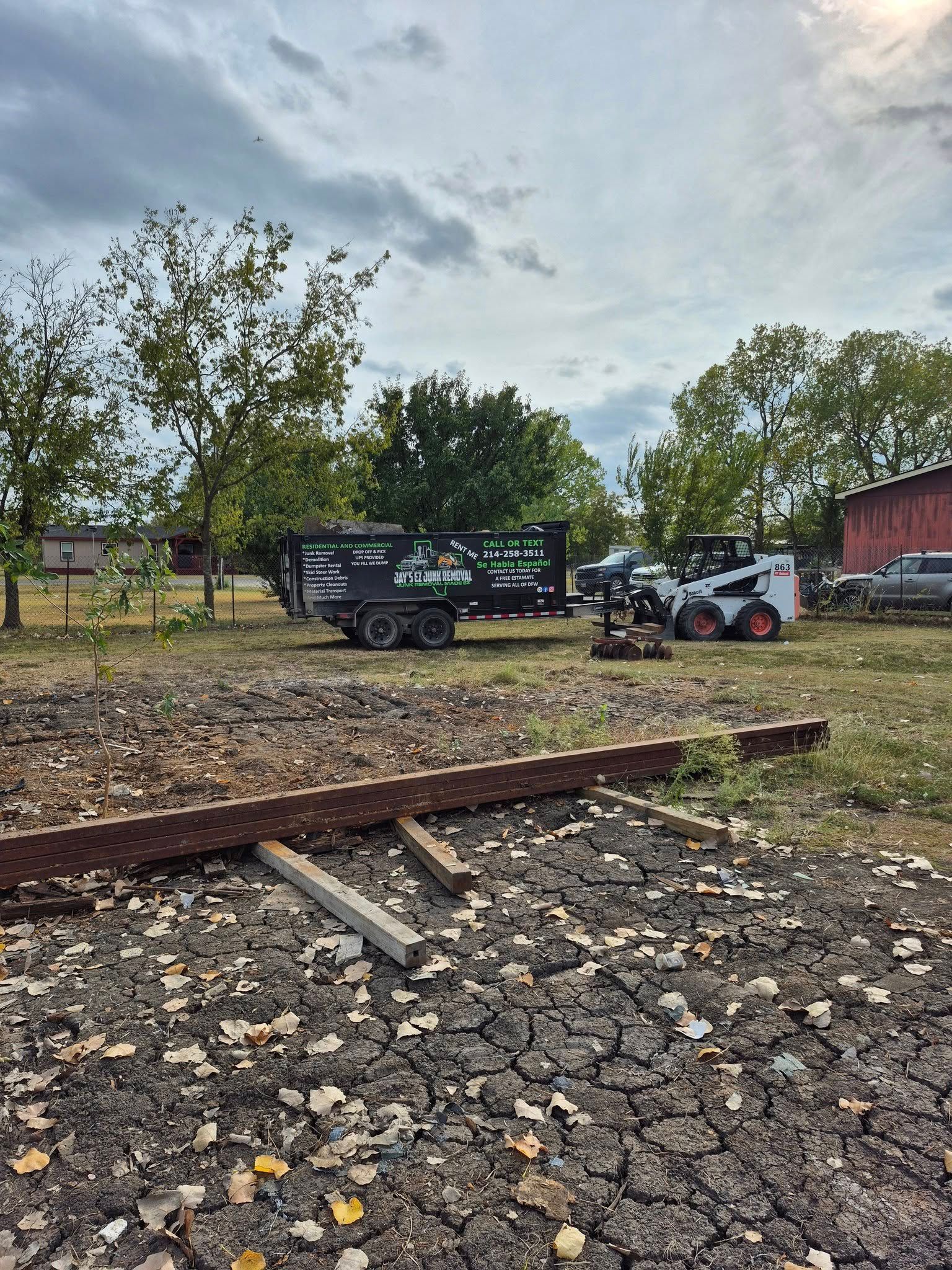 Construction site: Bobcat, trailer, and debris in a field with trees and a cloudy sky.