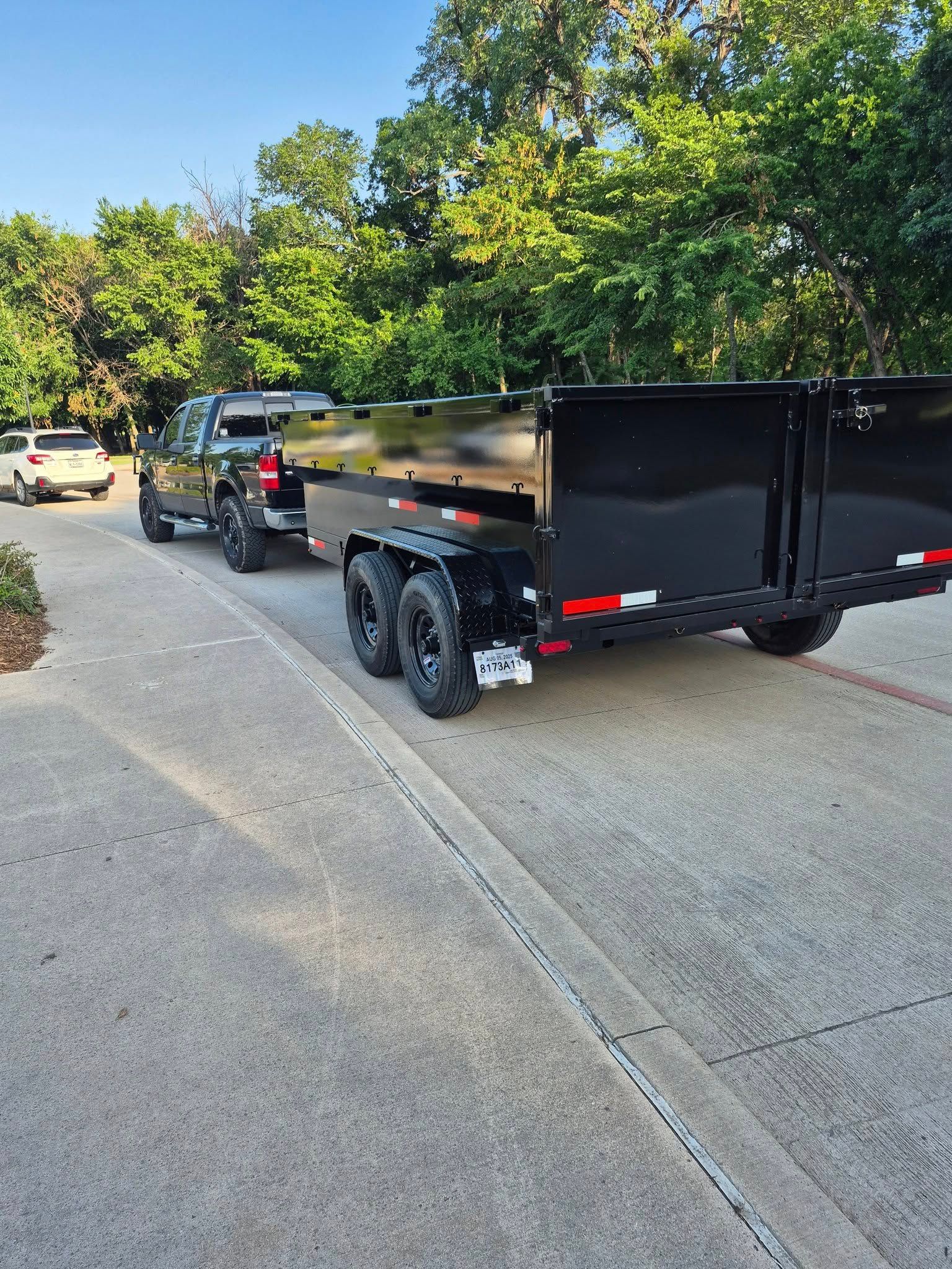 Black pickup truck towing a black dump trailer on a paved driveway, surrounded by greenery.