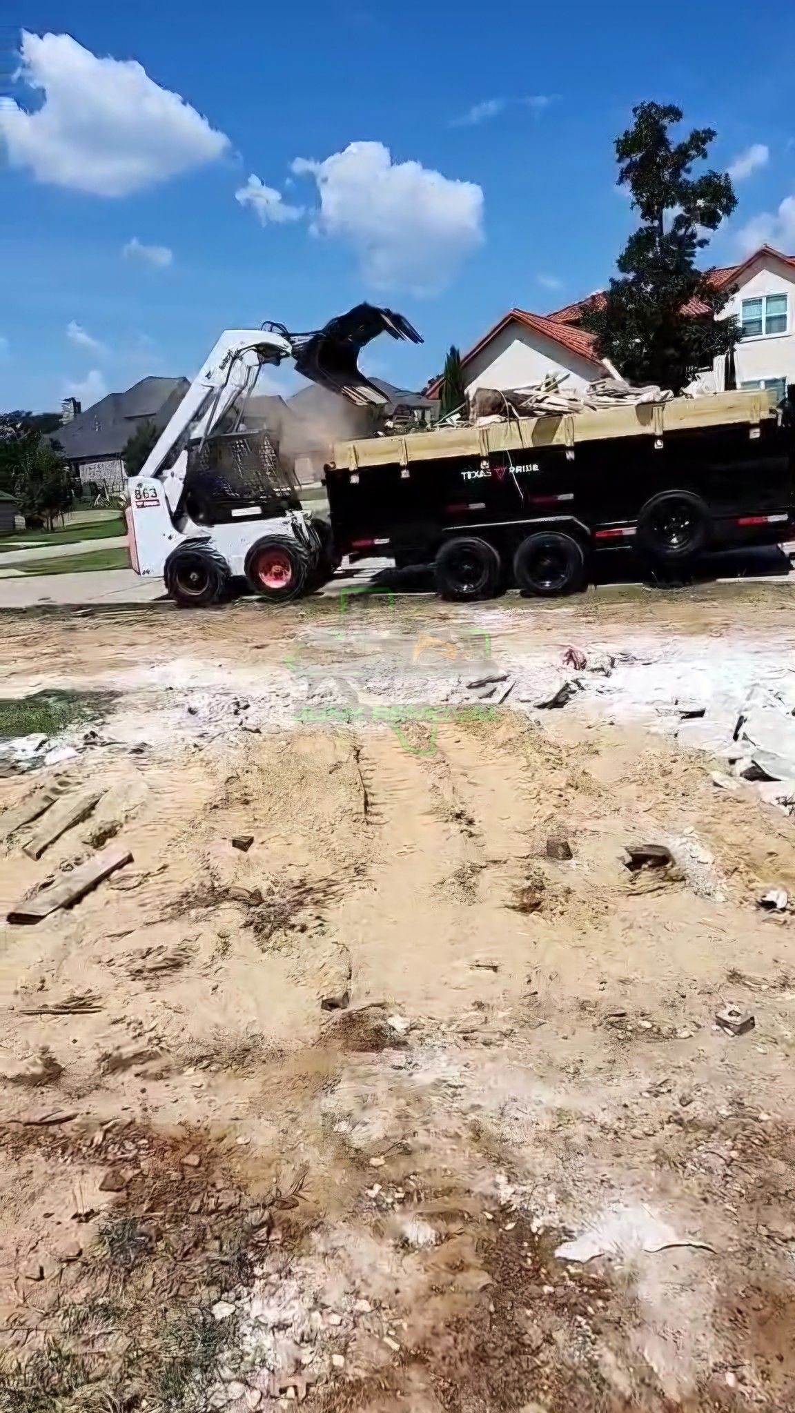 Bobcat skid-steer loader dumping debris into a dump truck on a construction site; houses in background.