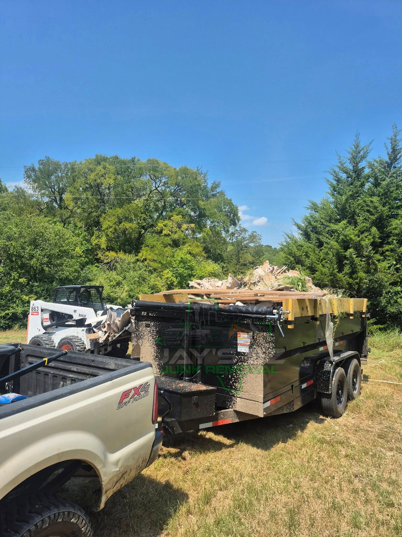 Truck pulling a trailer loaded with debris near trees under a blue sky. A Bobcat is beside the trailer.