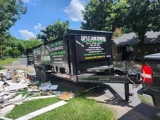 Dump trailer loaded with debris parked in a driveway. Green grass, trees, and blue sky in background.