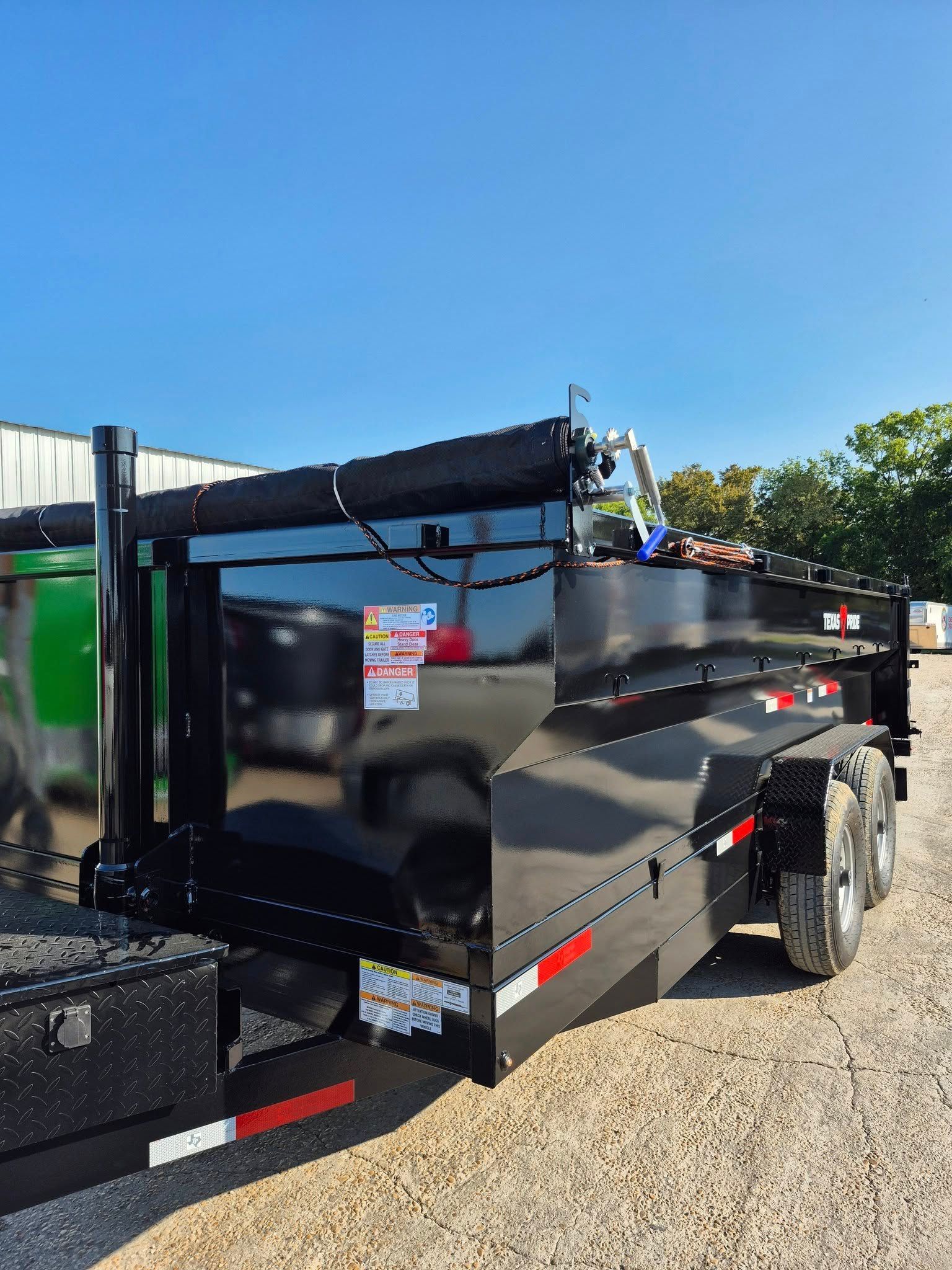Black dump trailer on gravel under a blue sky, near a green dumpster and building.